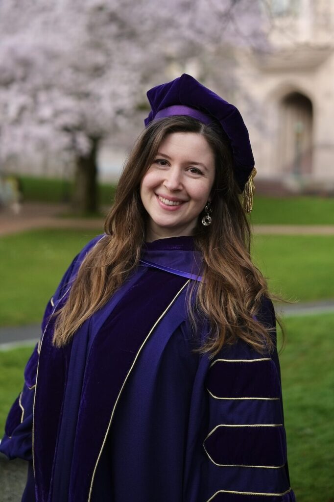 Gökçe-Alpkılıç wearing doctoral graduation regalia while standing in front of trees and campus building.